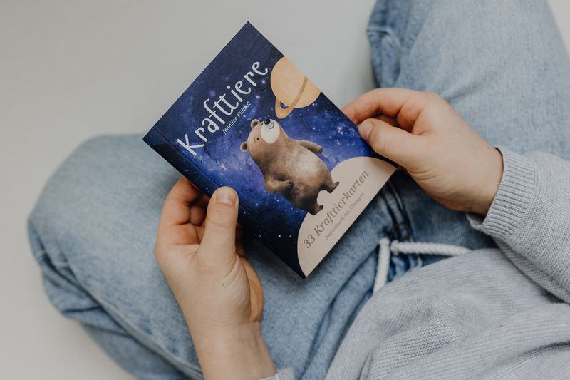 Ein Kind hält das Begleitbuch der Krafttierkarten von Jennifer Kühnel, das aus einer liebevoll gestalteten magnetischen Aufbewahrungsbox, 33 Krafttier-Affirmationskarten und einem Buch besteht. Das Begleitbuch fördert die spirituelle Verbindung zwischen Krafttieren und dem Körperbewusstsein, um Spannungen, Gefühle und Stress besser wahrzunehmen und abzubauen.  Ideal für Kinder und Eltern-perfekt  als Geschenk zu Ostern, Weihnachten, zur Einschulung oder zum Geburtstag.